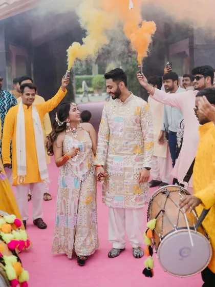 The couple makes a grand entrance to their Haldi ceremony, walking hand-in-hand through a cloud of yellow smoke bombs while a dhol player leads the way.