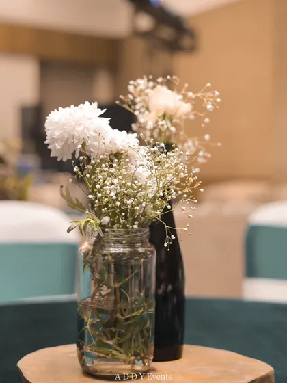 A minimalist centerpiece featuring white chrysanthemums and baby's breath in a simple glass jar and a dark bottle. Placed on a rustic wooden slice, this arrangement is perfect for adding a touch of understated charm.