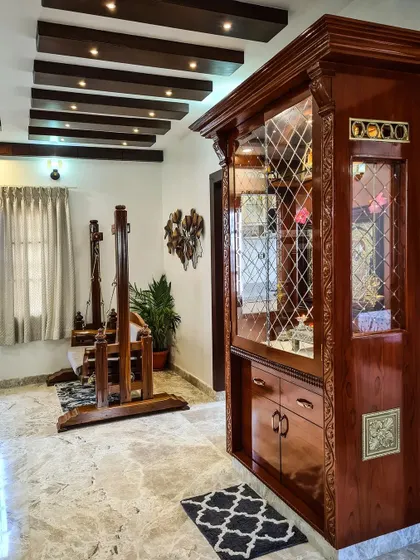 This image showcases a beautifully crafted pooja cabinet made from polished dark wood with detailed carvings and latticework on the glass doors. Positioned in the living area, it serves as both a spiritual corner and a piece of fine furniture.