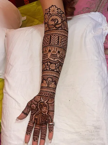 A full-arm bridal design featuring a groom's portrait on the palm. The arm is decorated with bands of traditional Rajasthani art, including peacocks and temple bells.
