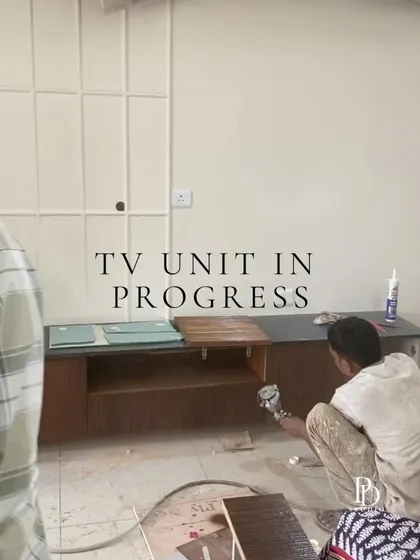 The TV unit is in progress. Here, a carpenter is working on the wooden console, with the wall paneling behind it already installed and awaiting its final coat of paint.