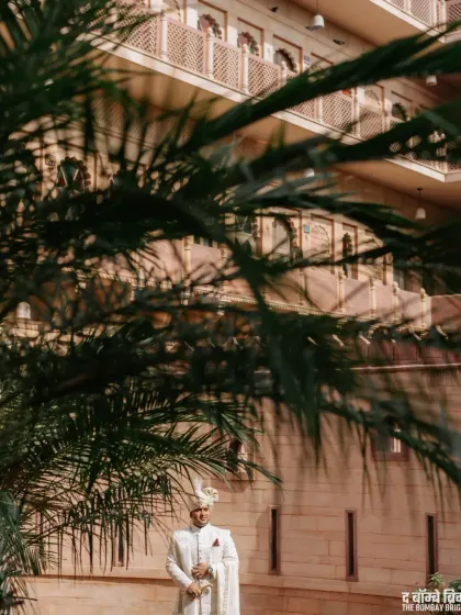 The groom looking regal in his white sherwani, framed by palm leaves at his palace wedding in Rajasthan.