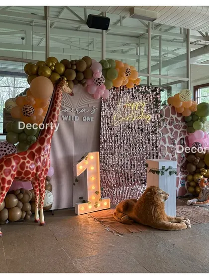 A chic "Wild One" jungle safari theme. The backdrop includes a rose gold sequin wall, a neon "Happy Birthday" sign, and life-sized giraffe and lion props for a glamorous touch.