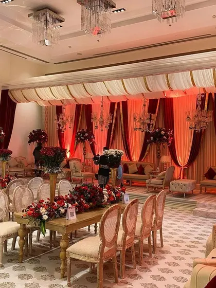 An indoor wedding setup that exudes classic elegance. Rich red and cream drapes frame the space, while crystal chandeliers and lush floral centerpieces create a warm, romantic, and traditionally grand atmosphere.