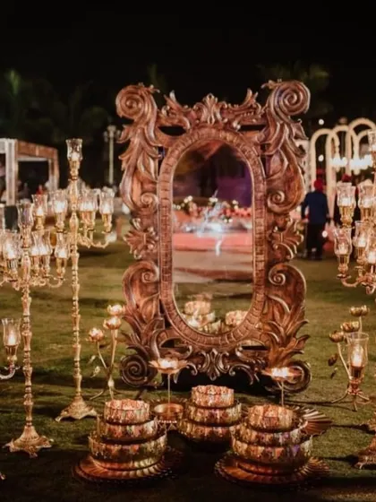 An ornate, vintage-style mirror surrounded by golden candelabras creates a stunning decor piece for an outdoor event. It adds a touch of royalty and old-world charm.