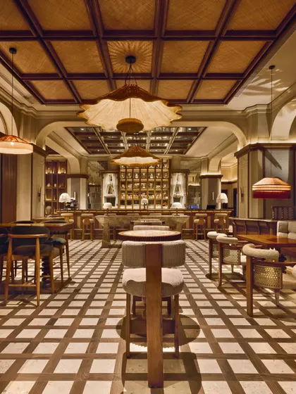 A wide view of the One8 Commune restaurant in Hyderabad, showcasing its tropical maximalist aesthetic. The space is defined by a symphony of teakwood, rattan light fixtures, and a patterned floor, creating a vibrant yet sophisticated atmosphere for dining.