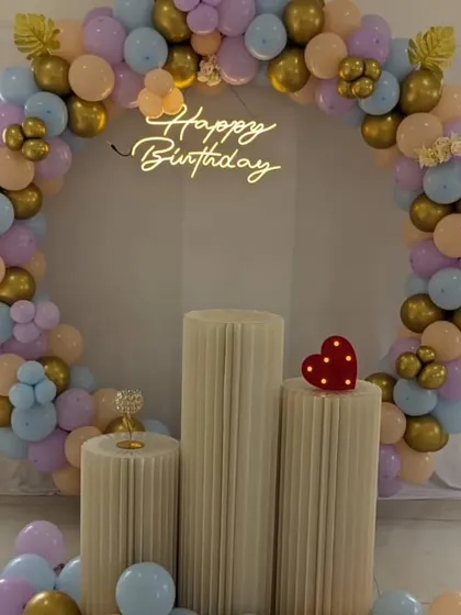 A beautiful birthday setup with a circular balloon arch in pastel and gold. The 'Happy Birthday' neon sign adds a modern touch to the decor.