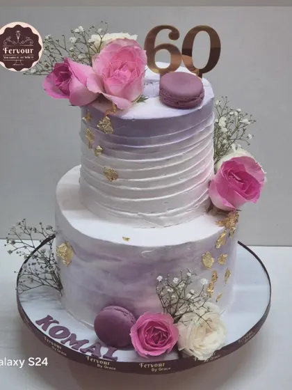 A beautiful two-tier cake for a 60th birthday, featuring a soft lavender watercolor effect. It's decorated with fresh roses, macarons, and a touch of gold leaf for an elegant and modern look.