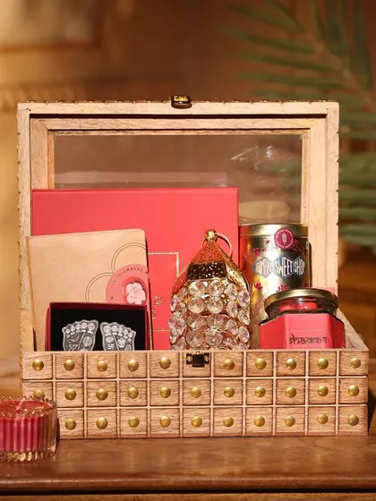 A stunning wooden box with brass stud detailing, filled with red-themed Diwali gifts like candles, sweets, and a decorative lantern.