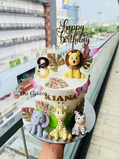 A sweet jungle animal cake for a third birthday, featuring a lion, elephant, giraffe, and zebra. The design is accented with butterflies and tropical leaves for a gentle, wild look.