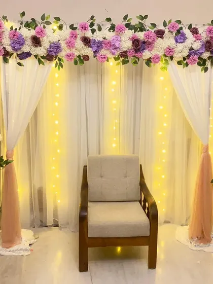 An elegant backdrop for a baby shower or naming ceremony. A floral arrangement sits atop a frame draped with white and peach fabric and fairy lights.