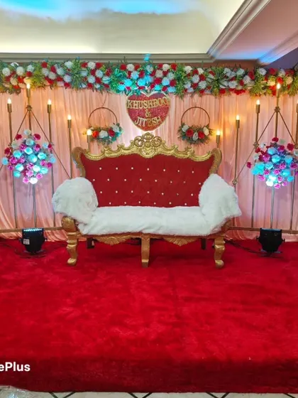 A classic engagement stage with a red and gold theme. The setup includes a beautiful red and gold sofa, floral arrangements, and a personalized heart sign with the couple's names, Khushboo and Jitesh.