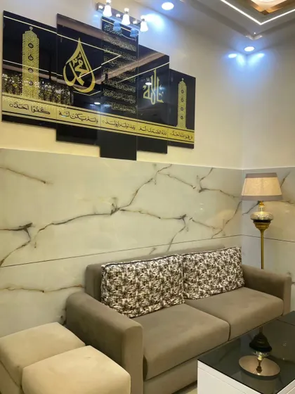 This living room features a comfortable sofa set against a wall with marble-finish panels. The large, elegant Islamic calligraphy wall art serves as a powerful spiritual and decorative centerpiece.