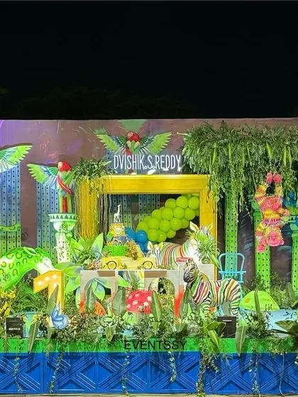 A vibrant neon jungle theme for a first birthday party. This wide shot shows the entire setup, filled with brightly colored animal props, lush greenery, and neon-style lighting effects.