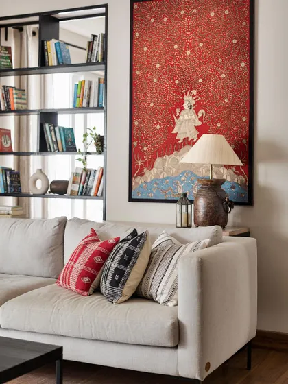 A large Pichwai painting adds a vibrant splash of color to the living room of the 'Ikiga Living' home. The neutral sofa and open bookshelf create a perfect gallery-like setting for displaying significant art pieces.