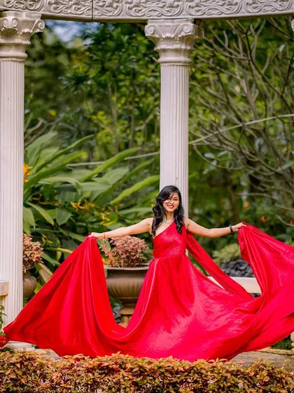 A happy and carefree moment in the garden. The bride-to-be holds out her flowing red gown, creating a beautiful fan shape against the lush greenery and white pillars.