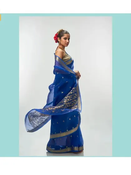 The magic of silk organza is in its weightless drape. This ethereal cerulean blue saree flows effortlessly, adorned with delicate gold zari buttis. The pallu showcases a rich floral motif, making it a piece of wearable art.