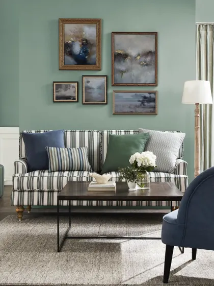 A classic living room with a striped sofa and a gallery wall of landscape paintings. The sage green walls and traditional furniture create a timeless, comfortable aesthetic.