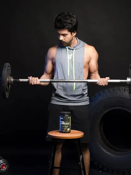 A male athlete performing barbell curls in front of a large tire, with a pre-workout supplement on a stool. The focus is on action and product association.
