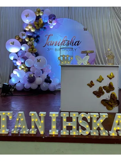 A beautiful butterfly themed third birthday setup with a purple and gold balloon arch, a personalized backdrop, and the child's name in large marquee letters.