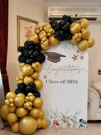 A graduation party decoration for the "Class of 2025". The setup includes an arched backdrop and a black and gold balloon garland.