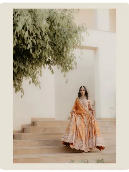 The bride descending the stairs, her lehenga in full view. The framing with the tree leaves adds a natural, soft touch to the portrait.