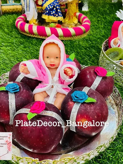 A baby shower platter featuring a doll in a pink robe, placed among decorated red apples. The silver ribbon and rose accents add a delicate touch.