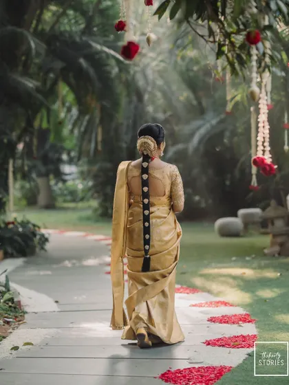 A graceful shot of the bride from behind, walking along a path strewn with flower petals. Her elegant hairstyle and the drape of her golden saree are beautifully featured.