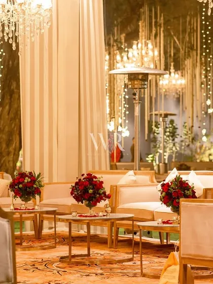 A regal lounge area with a backdrop of cascading mogra and chandeliers. The neutral-toned furniture is accented with deep red floral arrangements, tying it into the overall color scheme.