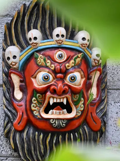 The Bhairava mask, framed by green leaves, showcasing its vibrant colors and protective symbolism. These hand-carved wooden masks are a significant part of Nepalese culture, designed to bring character to your walls.