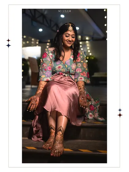 A full-length portrait of the bride at her Mehendi, showcasing her beautiful floral outfit and the intricate henna on her hands and feet. She sits with a serene smile, soaking in the moment.
