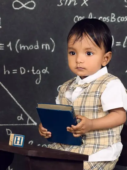 A baby boy in a plaid vest holds a book, looking thoughtful and studious at his little desk.