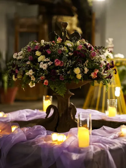 A romantic centerpiece that sets the mood. This arrangement of mixed florals in a vintage-style urn, surrounded by glowing candles on a bed of lavender fabric, creates an enchanting focal point for the table.