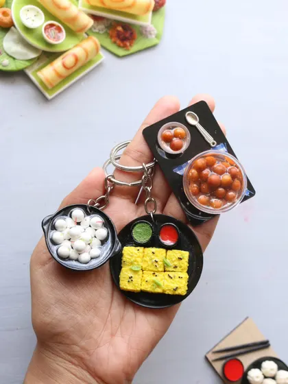 A hand holding a set of best-selling keychains: dhokla, gulab jamun, and kuzhi paniyaram.