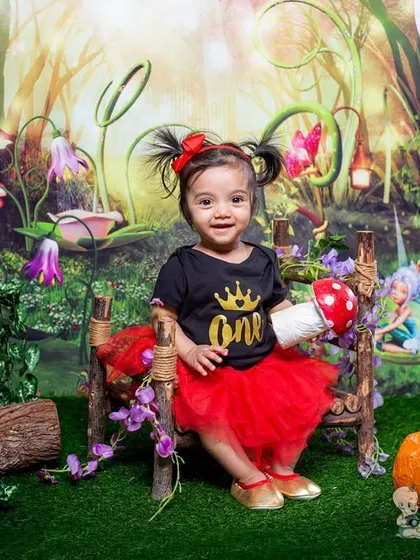 A little girl celebrating her first birthday in an enchanted forest. Dressed in a red tutu and a "one" shirt, she's the queen of this magical fairy-themed setup with mushrooms and pumpkins.