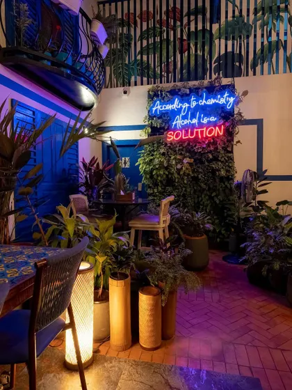 The Greek-themed courtyard transforms at night, with neon signs and ambient lighting creating a cool, vibrant atmosphere. The "Alcohol is a solution" sign adds a touch of humor and becomes a focal point.