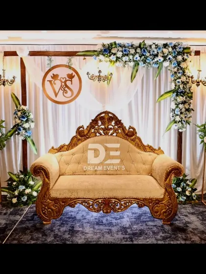 An elegant engagement stage featuring a custom monogram with the couple's initials. The design combines soft white drapery, a classic carved sofa, and asymmetrical blue and white floral arrangements for a sophisticated and personal look.