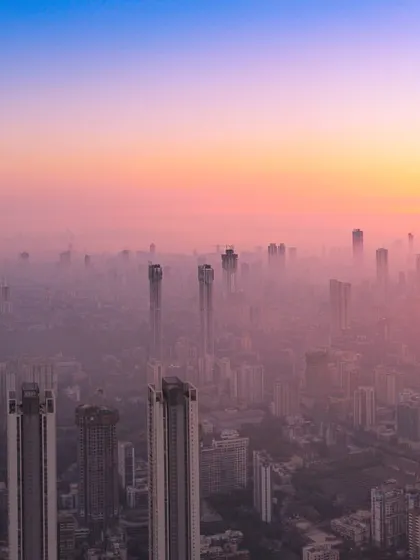 A panoramic drone shot of Mumbai's skyline enveloped in a thick layer of pink and yellow haze at sunset.