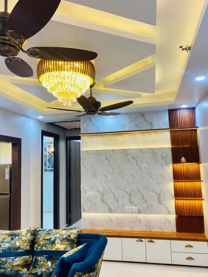 A beautiful drawing room featuring a statement TV wall with marble-finish panels and wooden fluted accents. The false ceiling design with a grand chandelier adds to the luxurious feel.