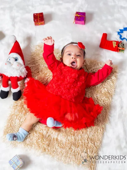 A baby's first Christmas! This little one in a festive red dress is surrounded by a Santa toy and presents, capturing the excitement and joy of the holiday season.