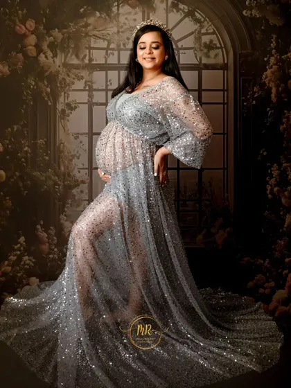 A glamorous solo maternity portrait. The mother-to-be glows in a sheer, sparkling silver gown, standing against a beautiful floral window backdrop in our studio.