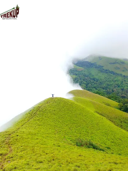 A lone trekker stands on a peak, dwarfed by the vastness of the green hills and clouds. It is a moment of pure solitude and connection with nature.