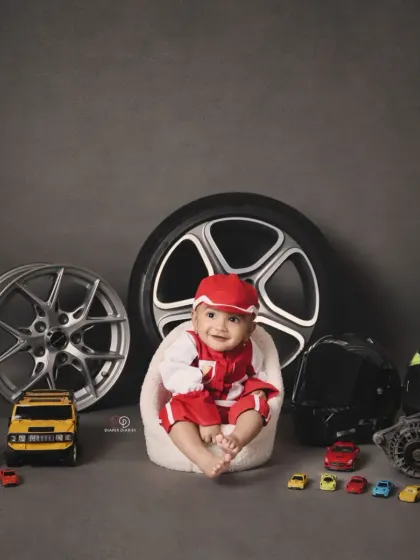 A full view of the 'Biker Baby' setup, featuring a baby boy in a red racing suit surrounded by real car parts, wheels, and toy cars. This is a great example of a highly customized themed session.