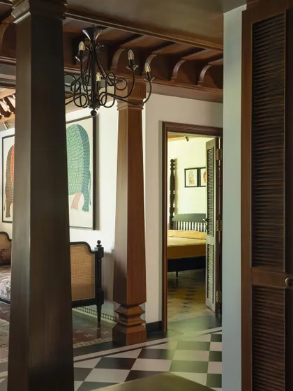 A glimpse from the main corridor into a bedroom, showing the continuity of the design language. The heavy teak pillars and checkered floor pattern flow seamlessly between spaces.