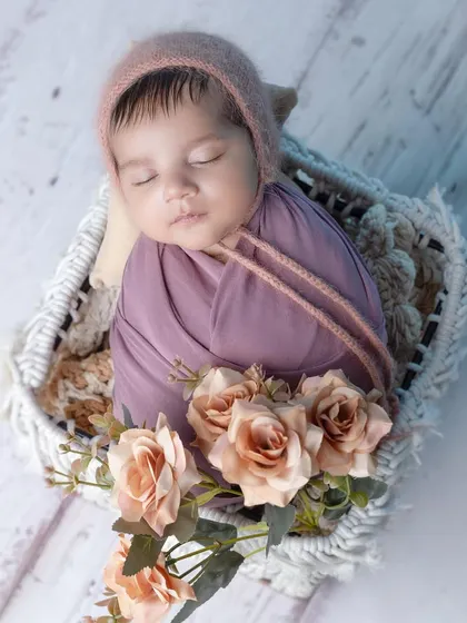 A sweet sleeping baby boy in a mauve wrap and bonnet, nestled in a white basket.