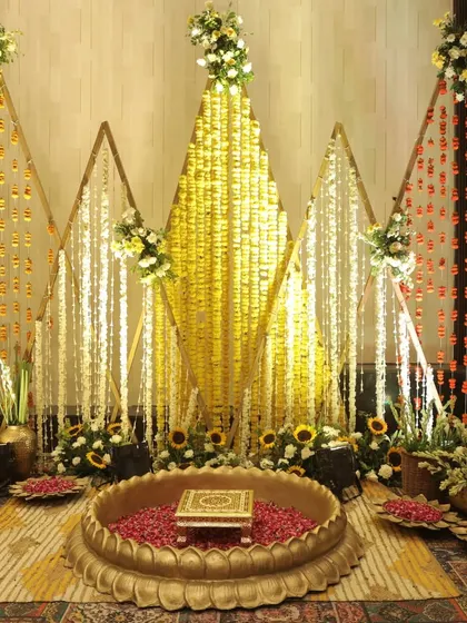 An elegant Haldi setup with triangular backdrops made of marigold strings in shades of yellow, orange, and white, centered around a traditional brass 'urli'.