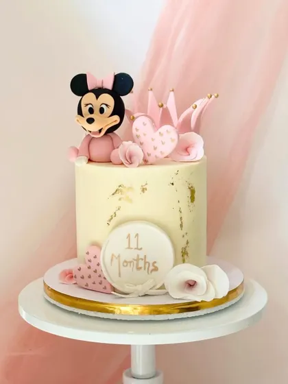A sweet Minnie Mouse cake for an 11-month celebration. It features a fondant Minnie figure, a pink crown, and delicate gold leaf accents on a smooth buttercream finish.