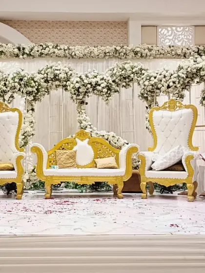 A regal stage setup for a reception, featuring ornate white and gold throne chairs and a backdrop overflowing with white flowers.