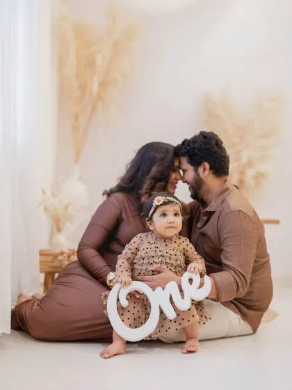 The birthday girl holds a "One" prop while her parents embrace lovingly behind her. This is a perfect family portrait to commemorate a first birthday milestone.