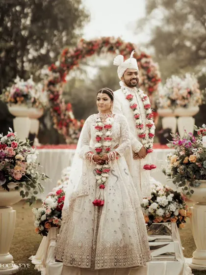 A formal portrait of the couple at their floral mandap, showcasing their coordinated white wedding attire and the beautiful decor.
